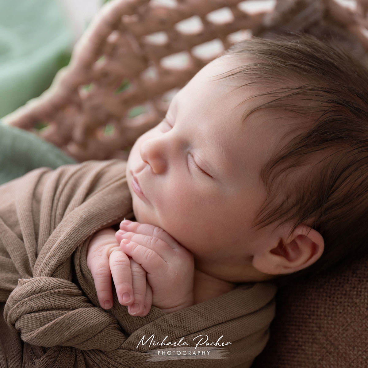 Seitliche Nahaufnahme eines schlafenden Neugeborenen, eingewickelt in braune Tücher, mit gefalteten Händen unter dem Kinn.
